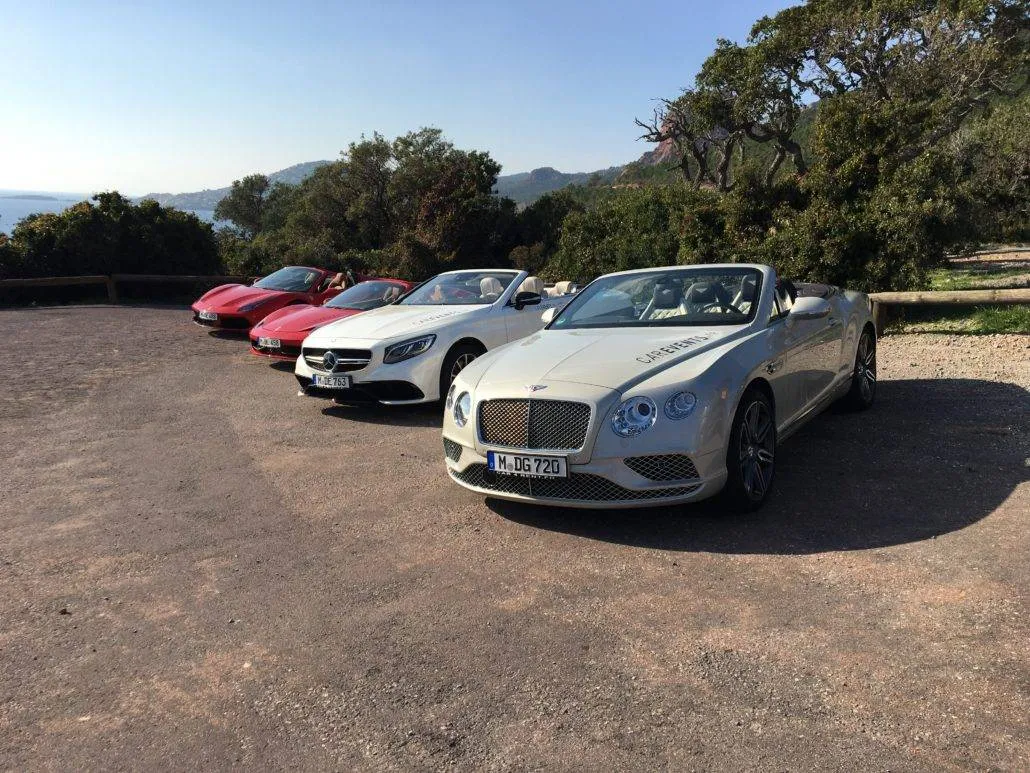 Luxury convertibles parked in scenic, secluded location with mountains and greenery in the background.