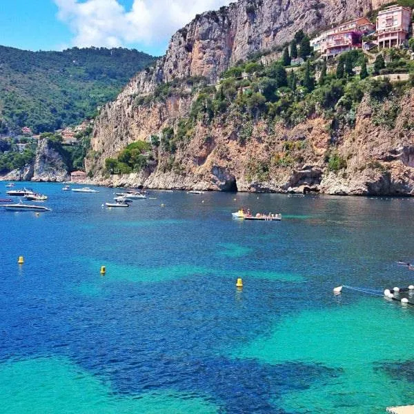 Turquoise bay with boats, cliffs, and red-roofed buildings under blue sky.
